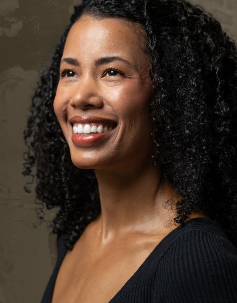 Smiling woman with natural curly hair looking upward, warmly lit against a textured neutral background.