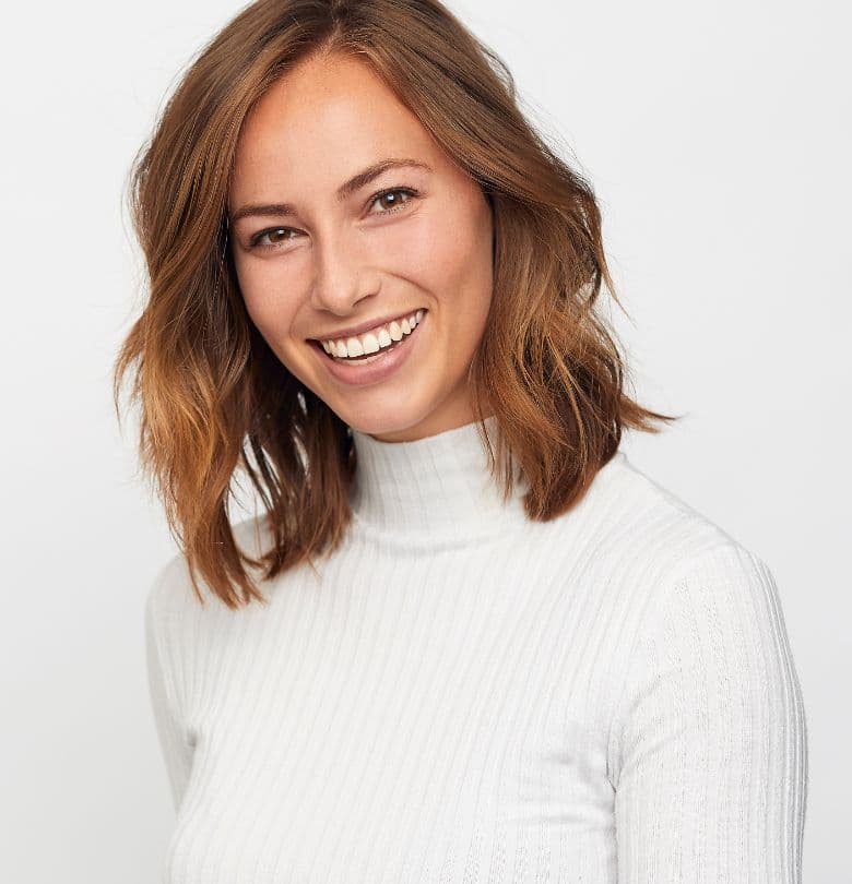 Smiling woman with shoulder-length brown hair wearing a white ribbed sweater against a clean white background, photographed in a bright, natural headshot style.