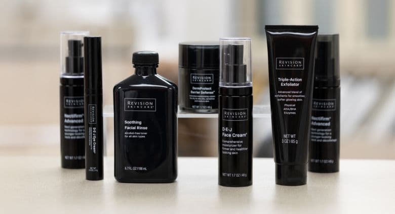 Lineup of black Revision Skincare bottles and tubes arranged on a light surface, showcasing multiple facial treatment and skincare products against a softly blurred background.